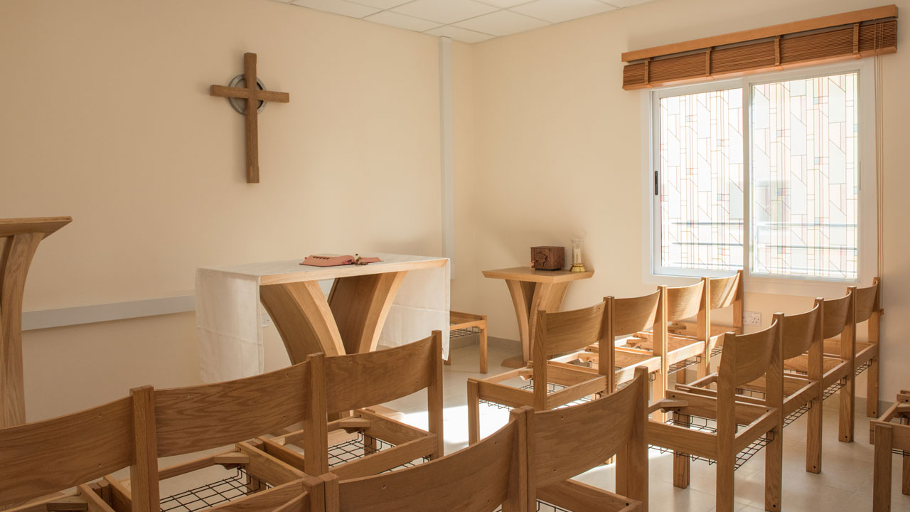 A small chapel with wooden chairs, an altar, and a cross on the wall, illuminated by natural light from a window