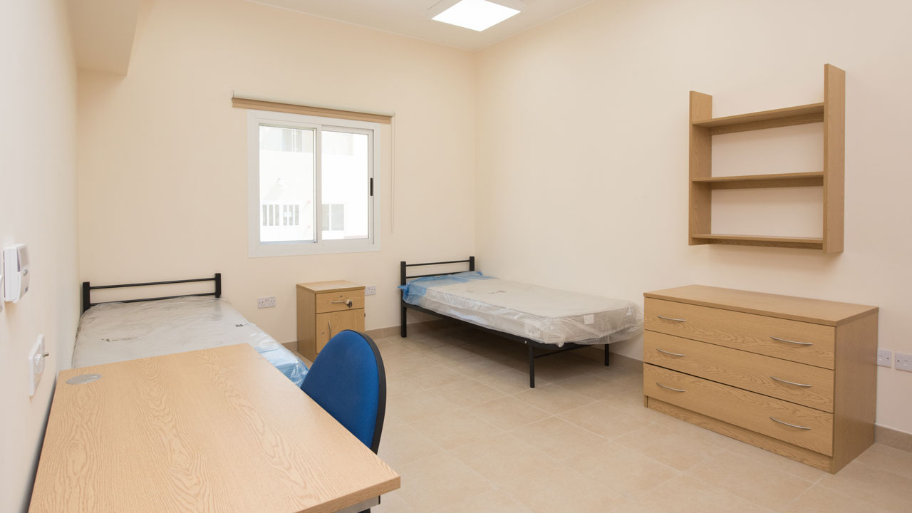 A simple bedroom with two beds, a desk, a chair, and a dresser, featuring a window that lets in natural light