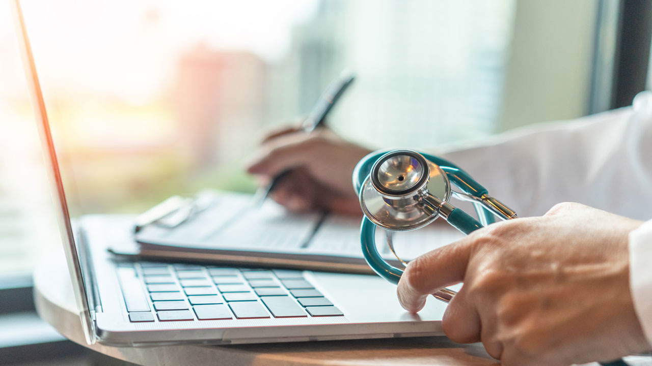 Person at a laptop with a pen in one hand, a stethoscope in another, and a laptop on the desk