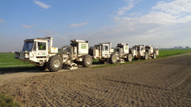 White trucks carrying surveying equipment, lined up at the edge of a field in a rural landscape