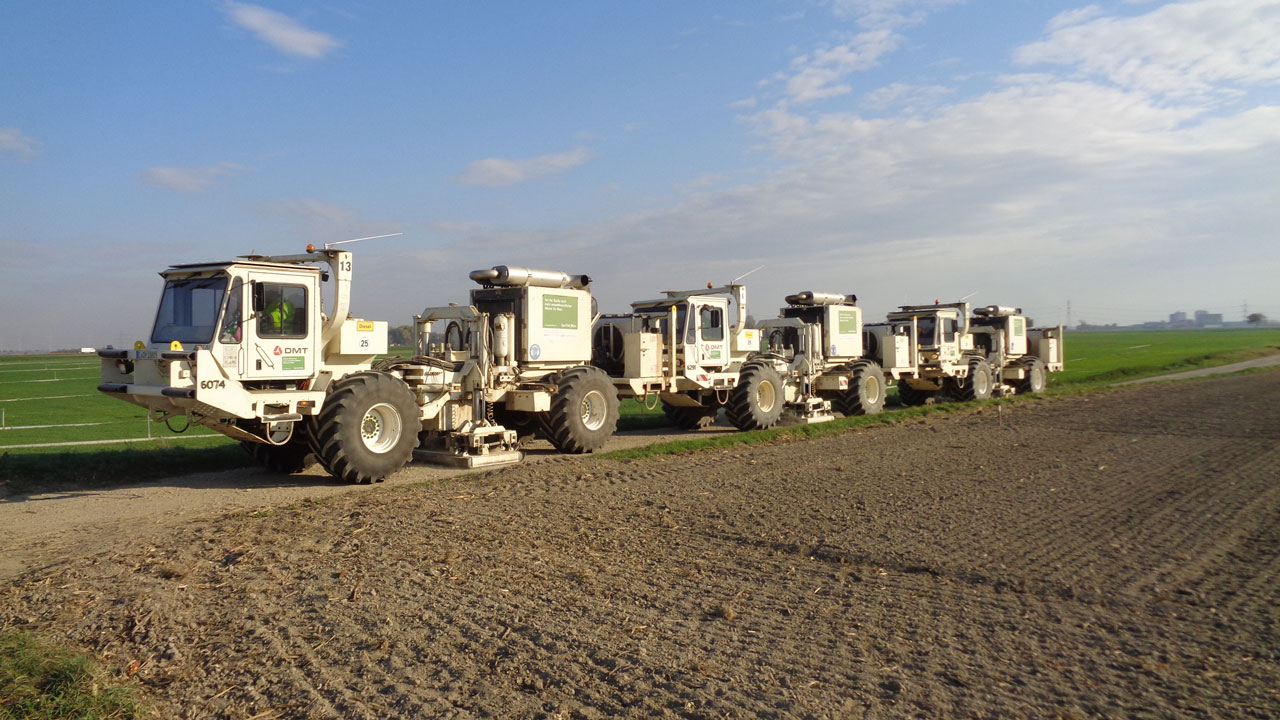 White trucks carrying surveying equipment, lined up at the edge of a field in a rural landscape