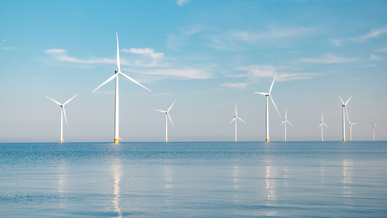Wind turbines on the horizon reflected in the sea, set against a pale blue sky