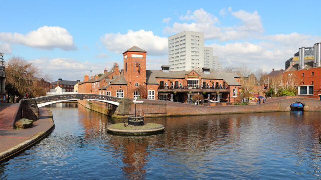 View across a canal in an urban setting with low rise redbrick residential buildings on the opposite bank, high rise residential blocks in the distance, and a footbridge crossing canal