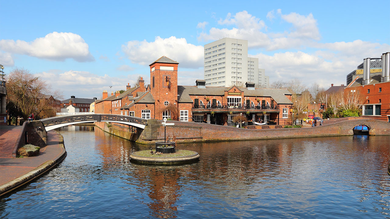 View across a canal in an urban setting with low rise redbrick residential buildings on the opposite bank, high rise residential blocks in the distance, and a footbridge crossing canal