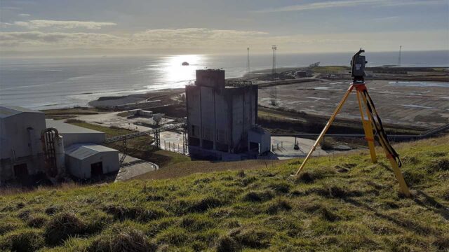 Wide lens view of the Aberthaw Power Station and geospatial surveying equipment on top of a hill with the sea in the background
