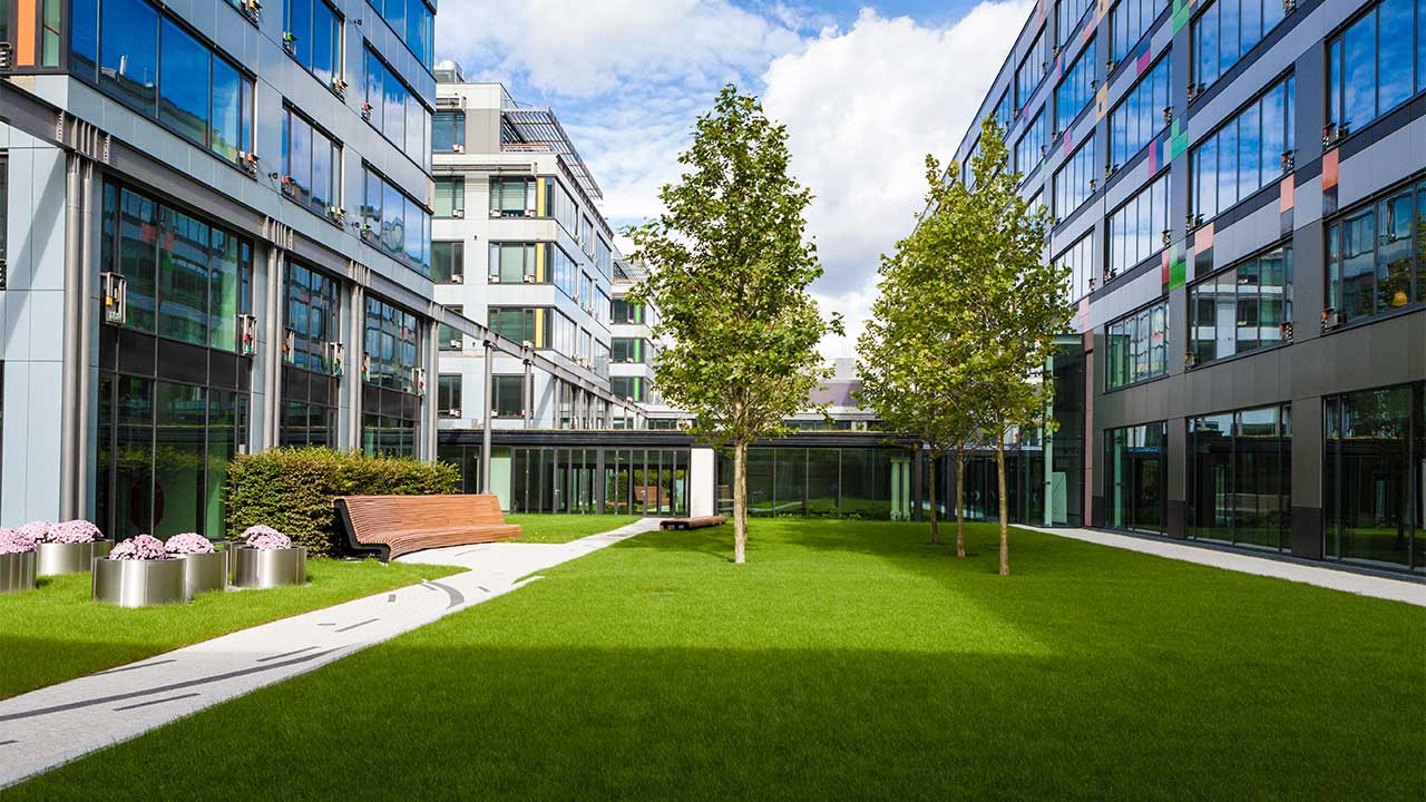 A courtyard area containing lawns and trees with a decoratively paved pathway and seating to one side, bordered on two sides by modern, glass-fronted, multistorey office buildings