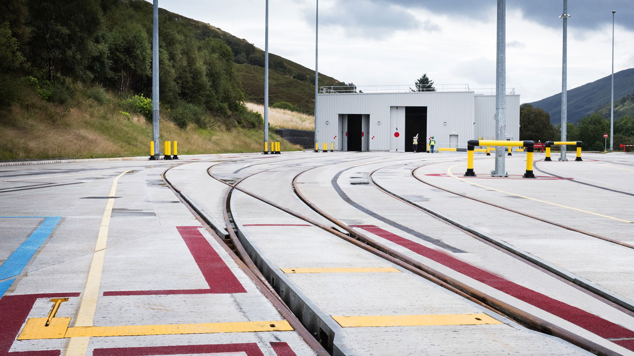 Curved railway tracks leading to a storage building, with safety barriers and light poles in a hilly landscape