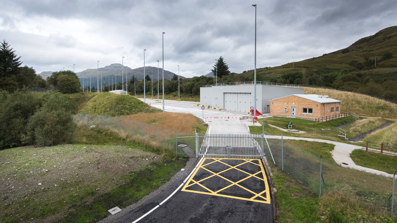 Entrance to a facility with a gated road, surrounded by greenery and hills, featuring storage buildings and tall light poles