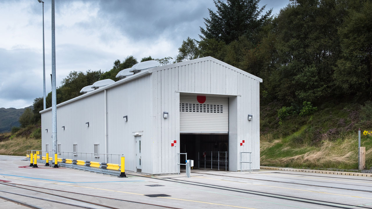 A gray storage building with a large roll-up door, surrounded by safety barriers and light poles