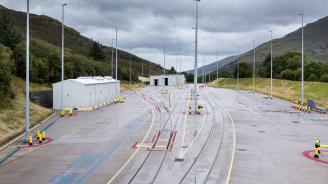 An expansive railway yard with multiple tracks, storage buildings, and surrounding hills under a cloudy sky