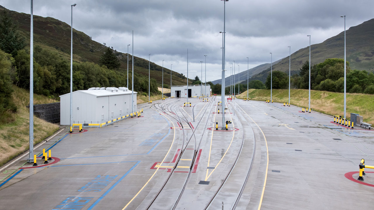 An expansive railway yard with multiple tracks, storage buildings, and surrounding hills under a cloudy sky