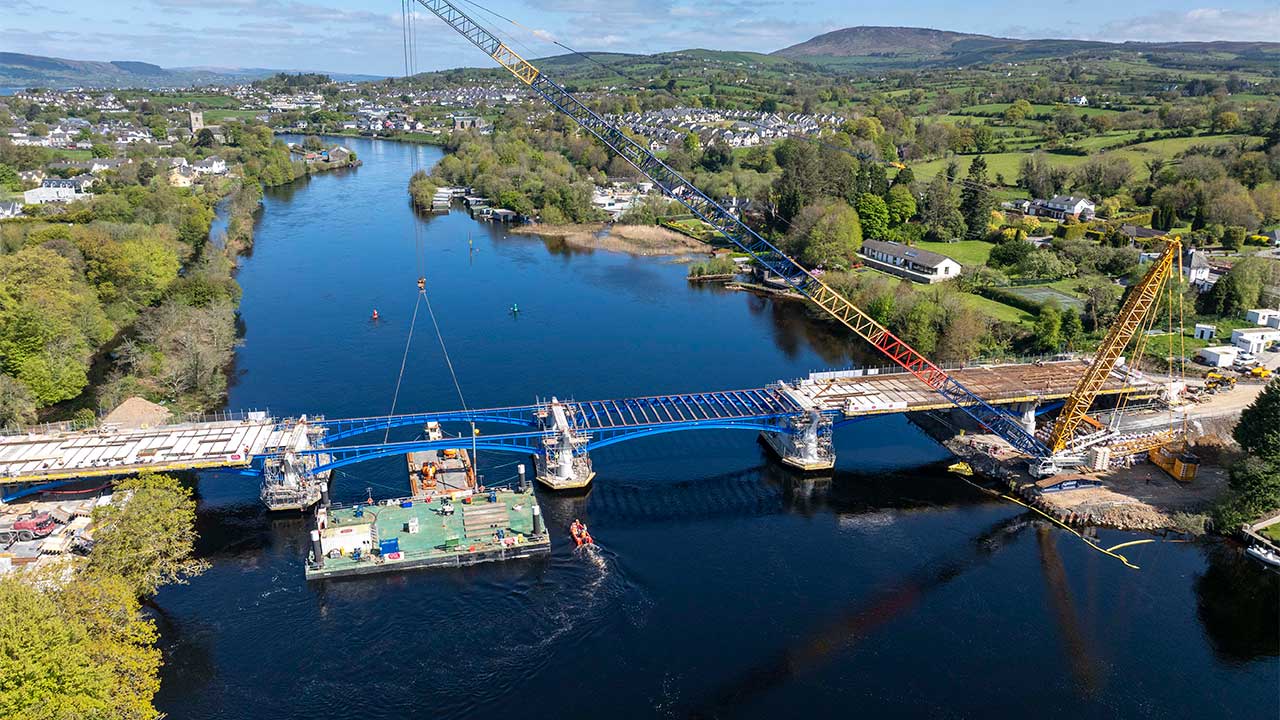 Aerial view of a road bridge under construction across a wide river, with a town visible on both river banks and green fields and hills in the background