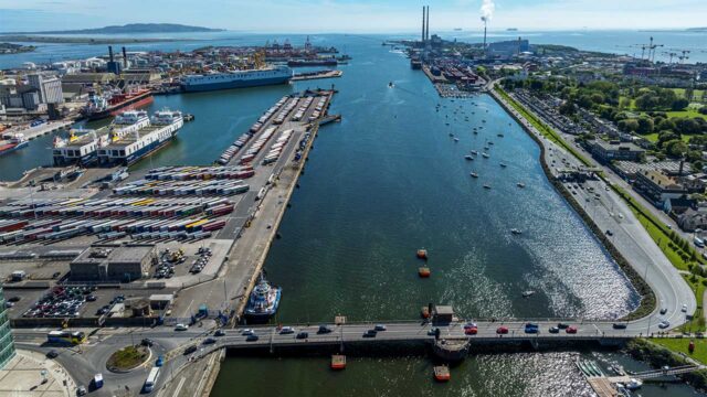 Aerial view of a commercial port showing containers lined up on the dockside, container ships in the loading berths, industrial buildings on the far side of the port with the sea in the distance and ships on the horizon