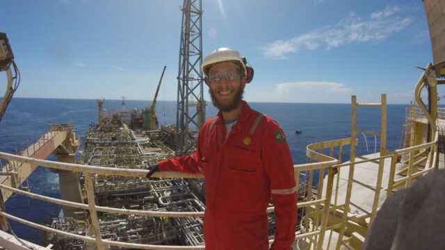Engineer in hard hat stands on a high platform with oil drilling rig visible behind