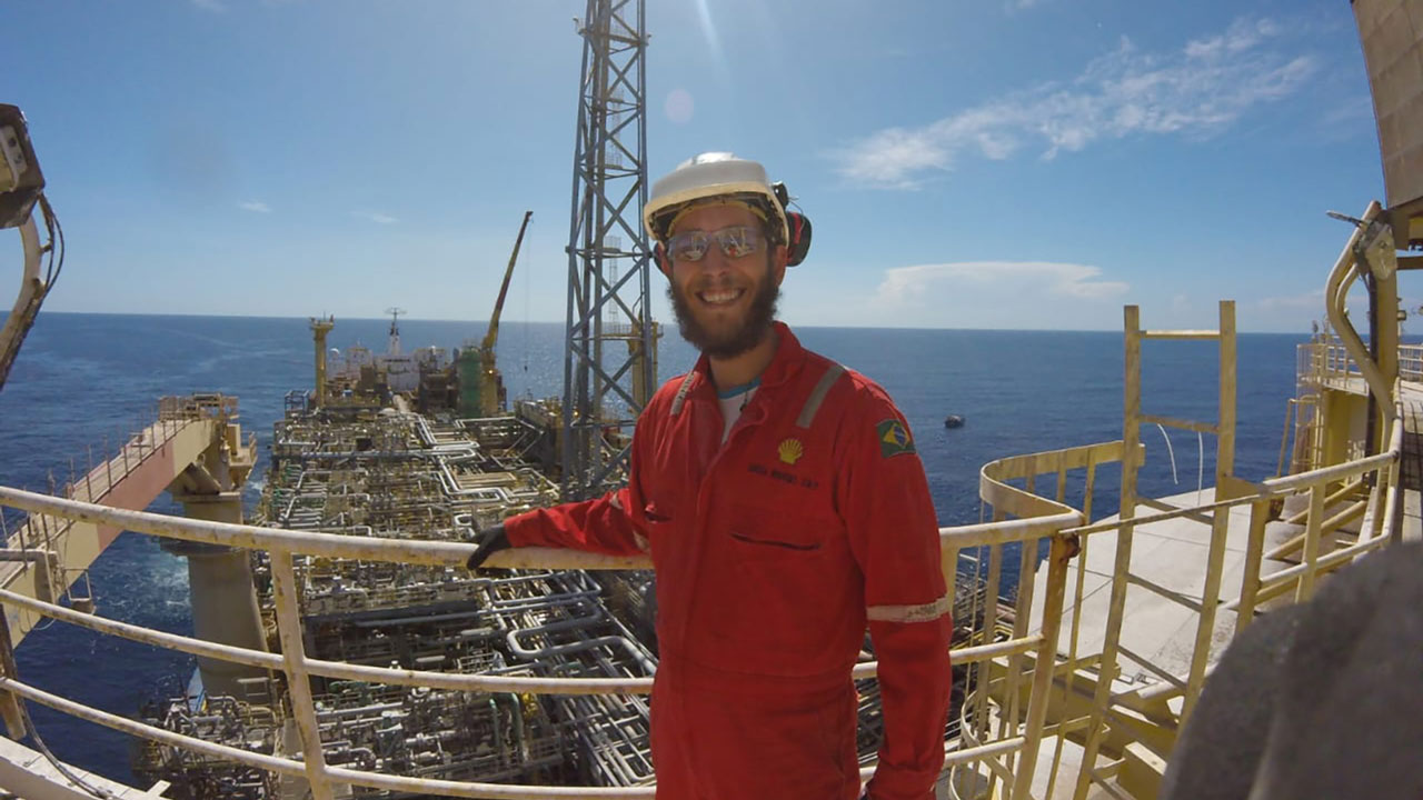 Engineer in hard hat stands on a high platform with oil drilling rig visible behind