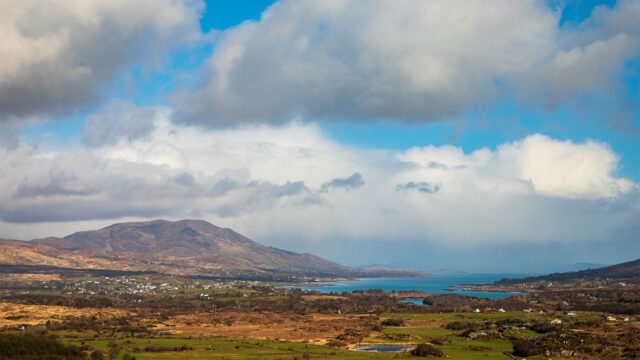 A view of a rural landscape looking across a wide sea inlet with mountains in the distance and a cloud-scattered blue sky