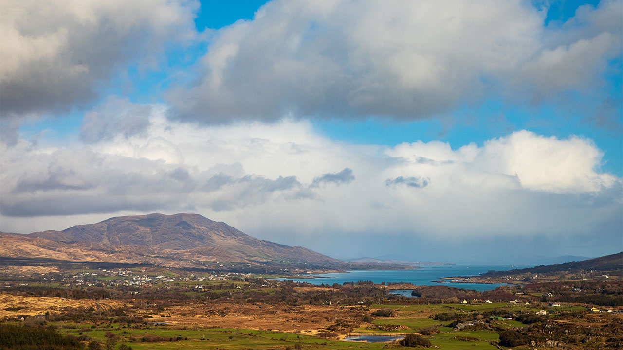 A view of a rural landscape looking across a wide sea inlet with mountains in the distance and a cloud-scattered blue sky