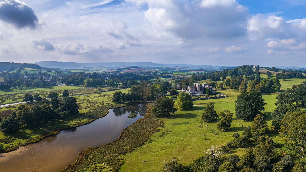 An aerial view across a rural landscape with an ornamental lake and historic country house in the foreground, and green fields and hills in the distance