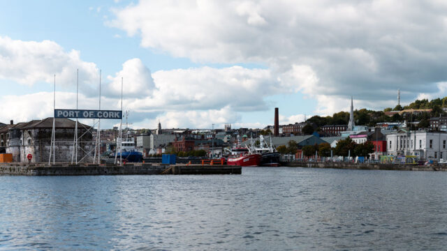 View across a commercial container port with several ships in dock , commercial buildings lining the port side and a town visible behind the port