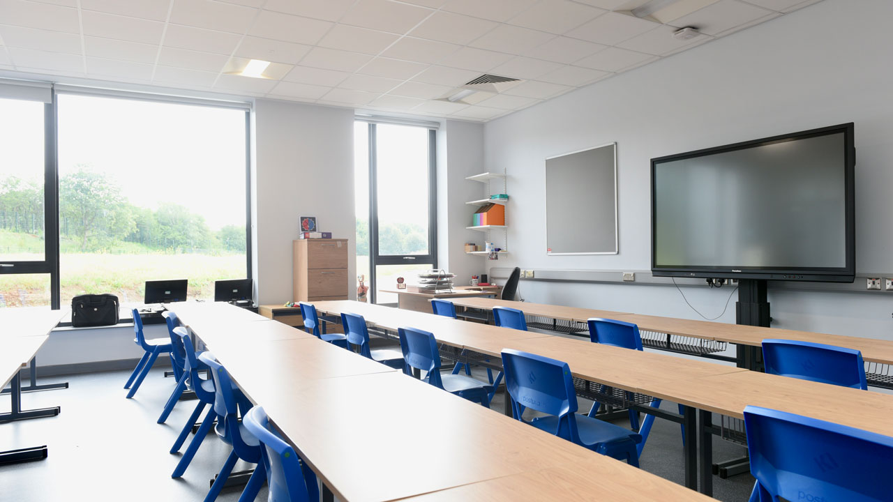 Interior of an empty classroom furnished with long tables, blue chairs, a screen at the front, and prominent windows letting in natural light