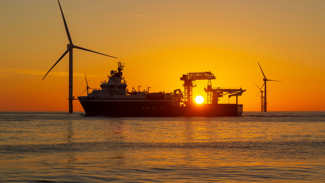 A ship on the sea against a sunset with offshore wind turbines on the horizon