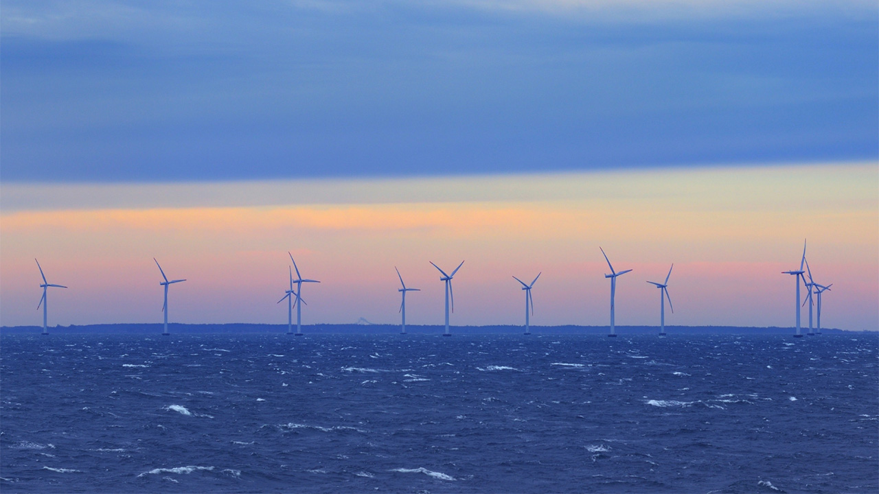 Multiple offshore wind turbines against the horizon with sea in the foreground at sunset