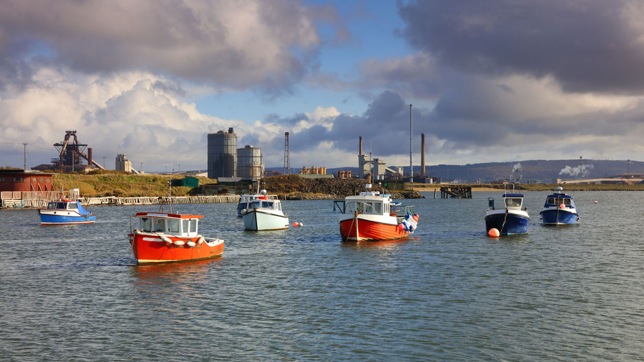 A line of moored fishing boats with industrial buildings in the background on the shoreline