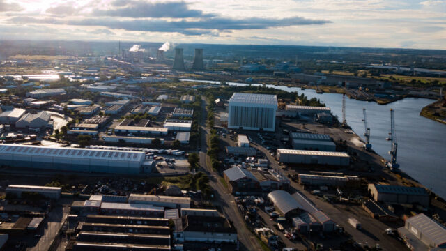 An aerial view of industrial buildings alongside a river with cooling towers of a power plant in the distance