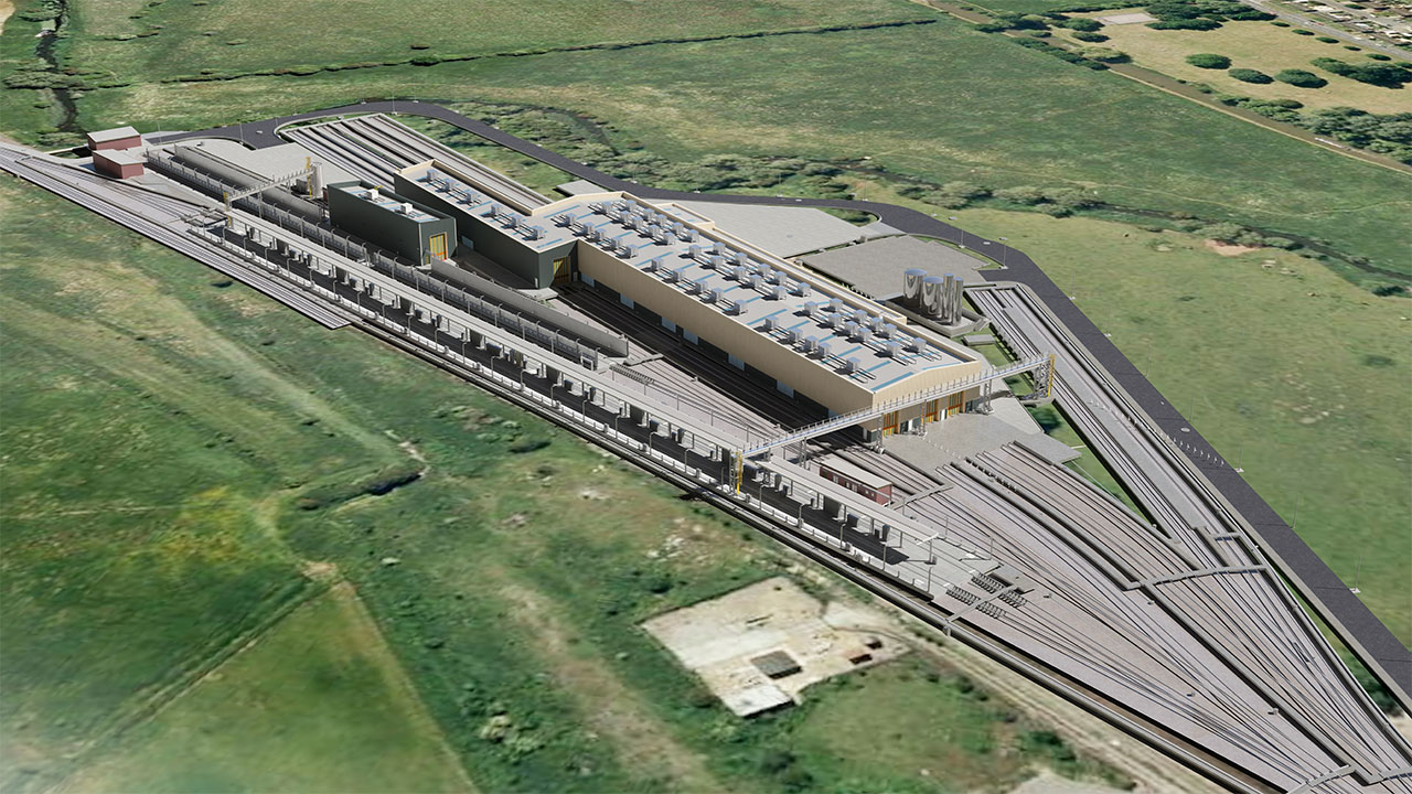 Aerial view of train station looking along train tracks into the station and station buildings, surrounded by green fields