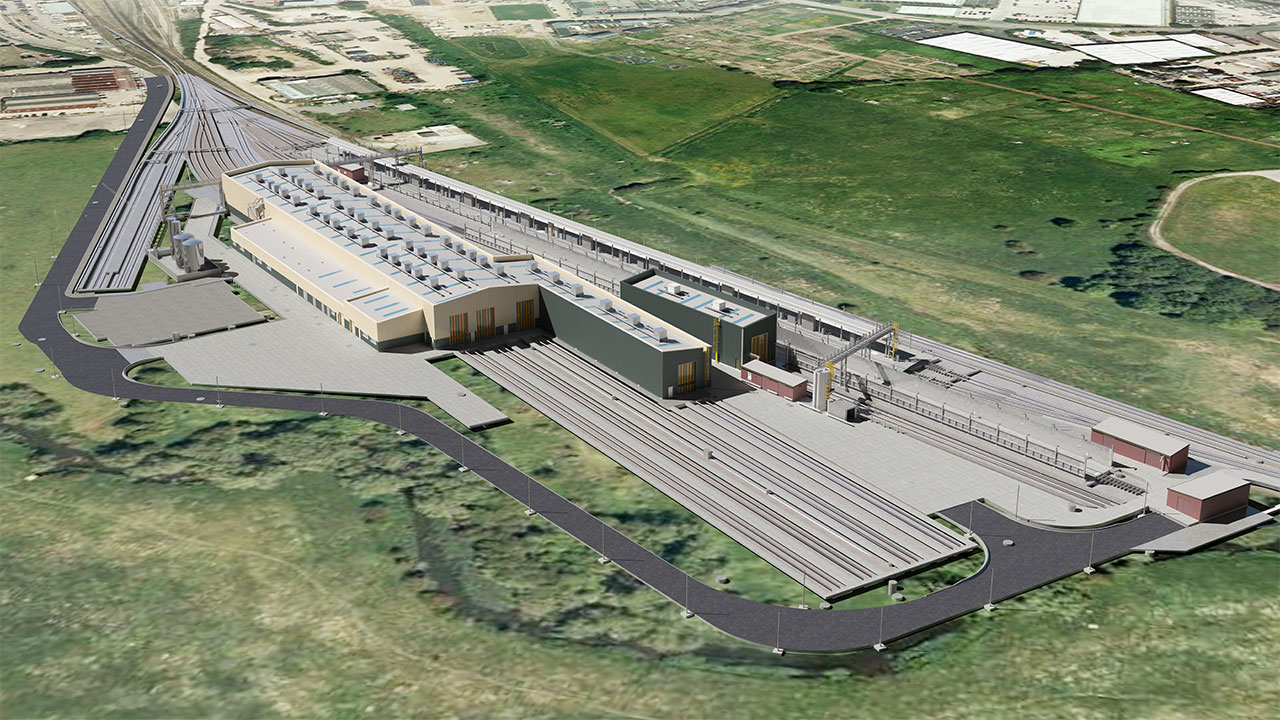 Aerial view of train station looking along train tracks into the station and station buildings, surrounded by green fields