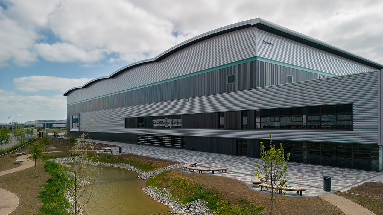 Side facade of modern, industrial building painted grey with trees, a small pond, seating and decorative paving in the foreground.
