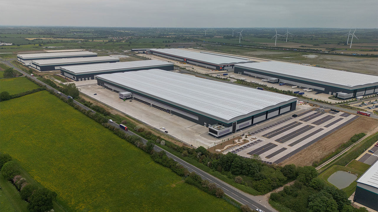 Aerial view of six large warehouse buildings set in a rural landscape with wind turbines in the distance