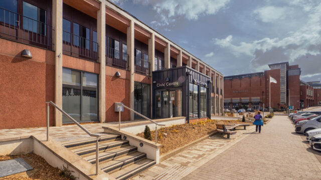 Image of a building with a set of steps at the front and a car park to the right