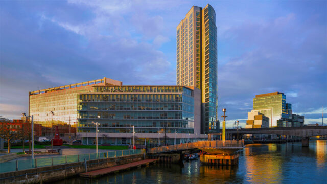 A view across a river towards high rise, glass fronted commercial offices, with a bridge to one side and grassed area alongside the river