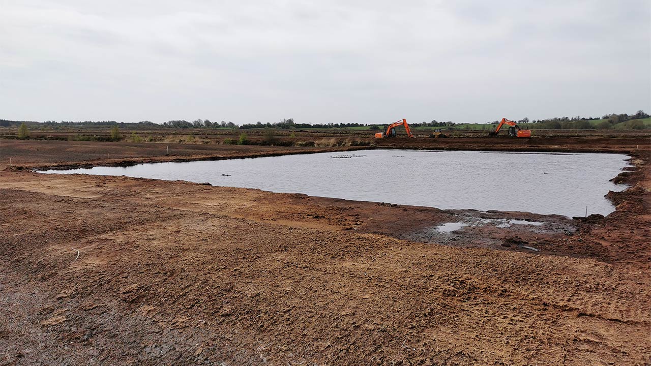 A large area of brown peatland with a pool of water in the foreground and excavators working in the distance