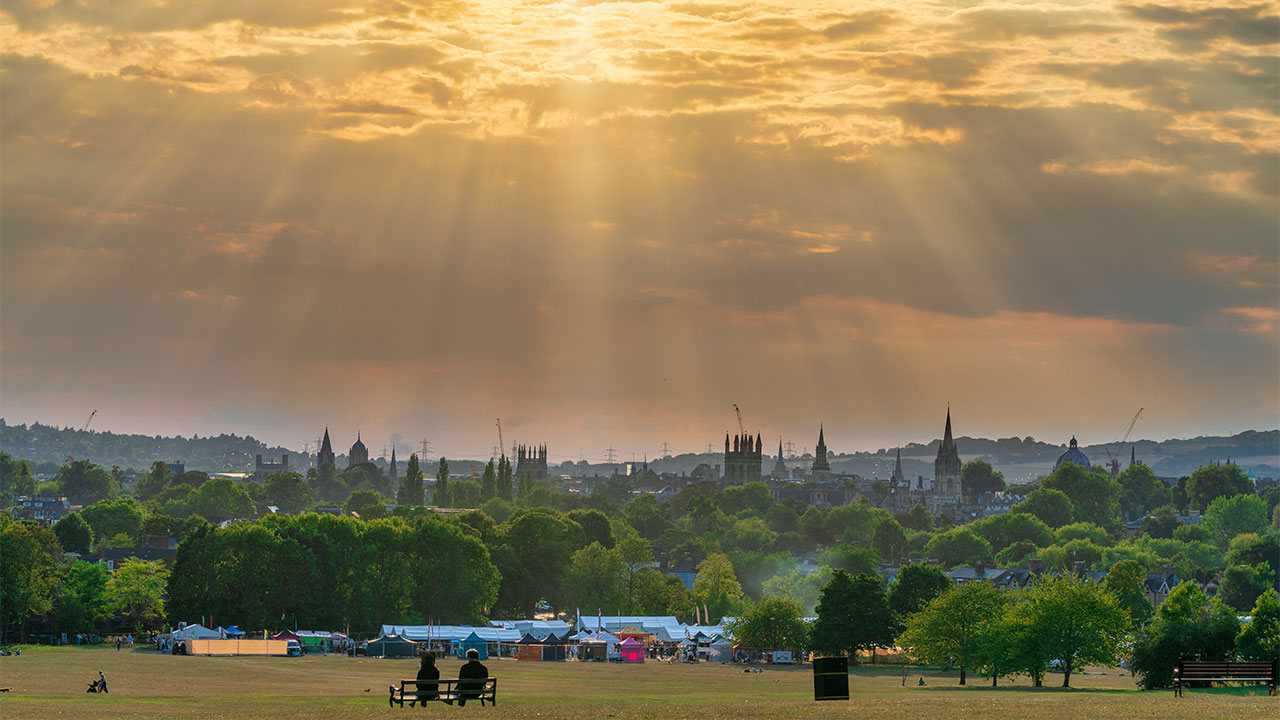 A view across parkland and trees to a city skyline with gothic towers and church spires, lit by shafts of sunlight breaking through storm clouds