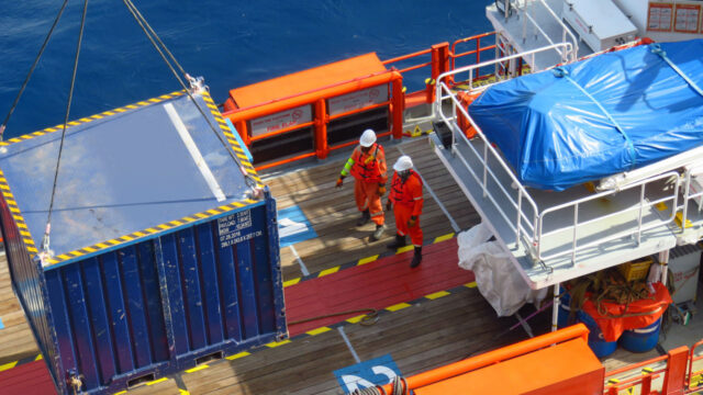 An aerial view of a shipping container being lifted from a ship’s deck by a crane with two crew members standing on deck