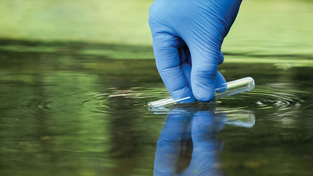 A gloved hand taking a sample of water with a test tube