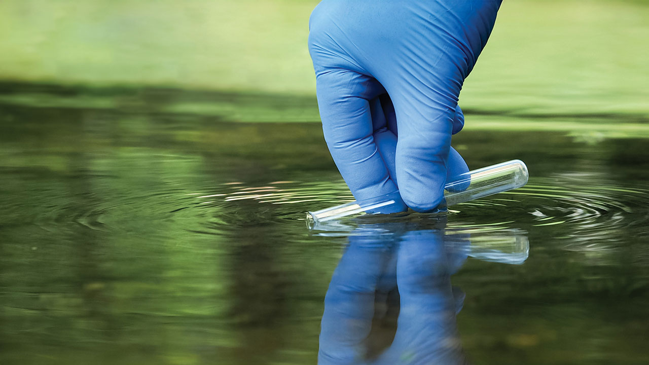 A gloved hand taking a sample of water with a test tube
