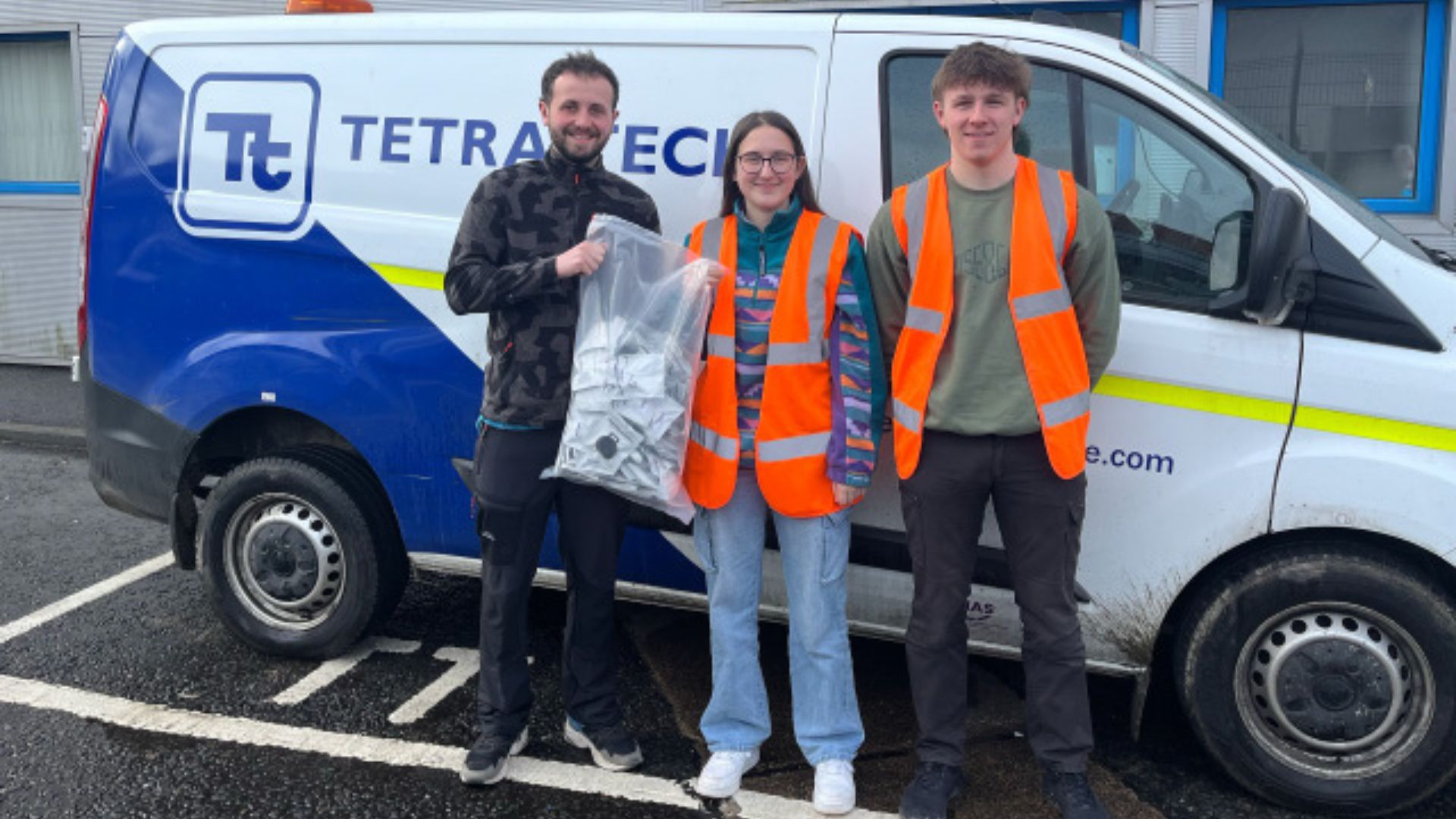 Three individuals stand in front of a Tetra Tech van, holding a bag of equipment