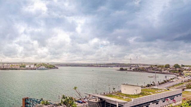 A view across a sea inlet with residential areas on the shoreline in the foreground, ships in a port area with loading cranes in the distance and green fields and hills on the horizon