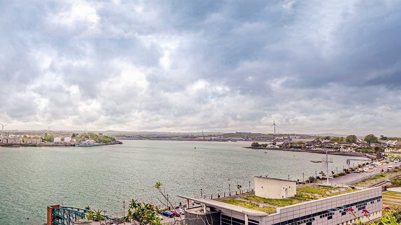 A view across a sea inlet with residential areas on the shoreline in the foreground, ships in a port area with loading cranes in the distance and green fields and hills on the horizon