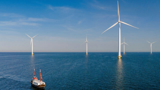 A fishing boat sailing alongside wind turbines on a calm sea against a blue sky