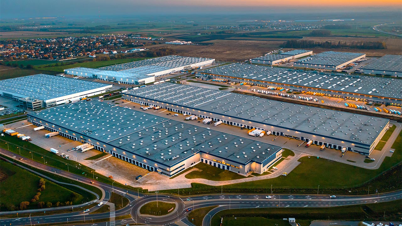 An aerial view of multiple warehouse buildings with a town in the background, access roads in the foreground and a sunset on the horizon