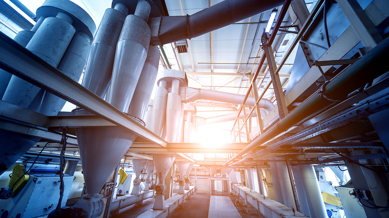 Interior of industrial processing plant with tall cyclone separators, interconnected ducts and piping, walkways, and bright sunlight filtering through ceiling