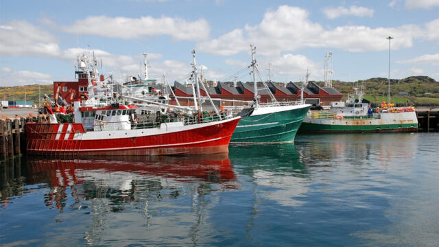 A view of three colourful fishing boats moored at the harbourside with modern buildings behind and green hills in the distance