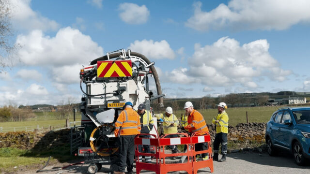 Group of workers at the side of a road with machinery behind them and a field and a cloudy sky in the background