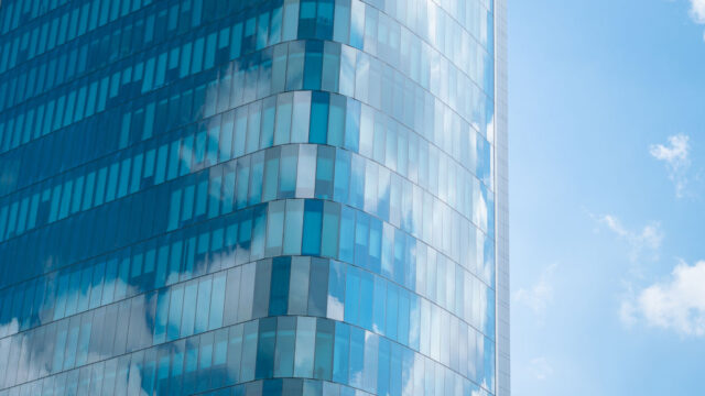 A high rise building against a blue sky with a reflection of buildings in the glass