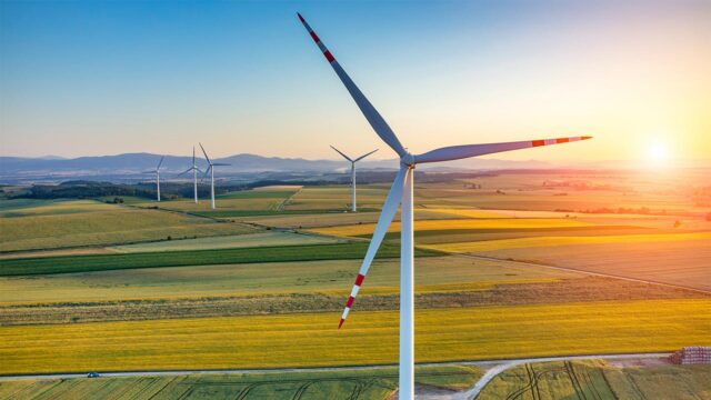 Close-up wind turbine overlooking patchwork farmland at sunset, distant turbines and rolling hills under a golden sky