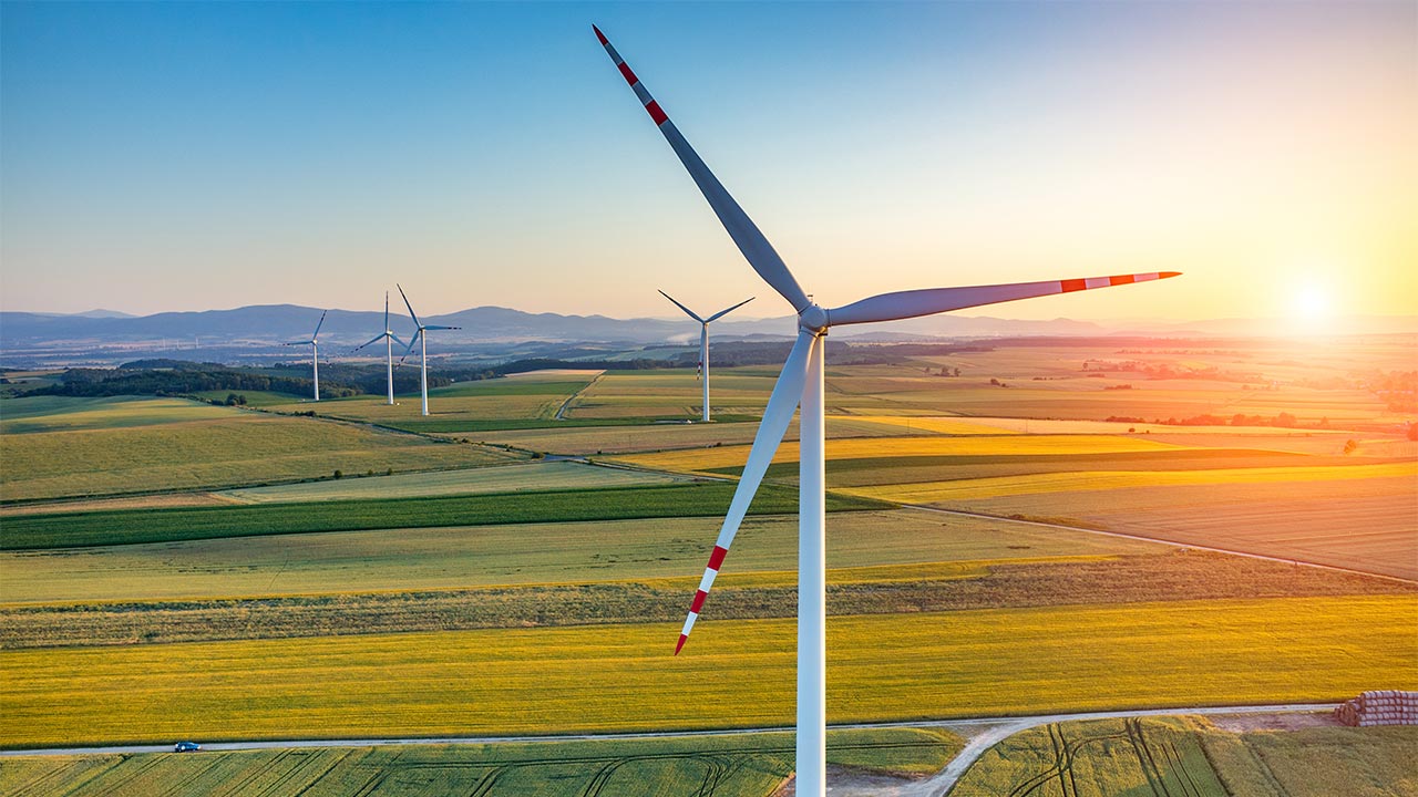 Close-up wind turbine overlooking patchwork farmland at sunset, distant turbines and rolling hills under a golden sky